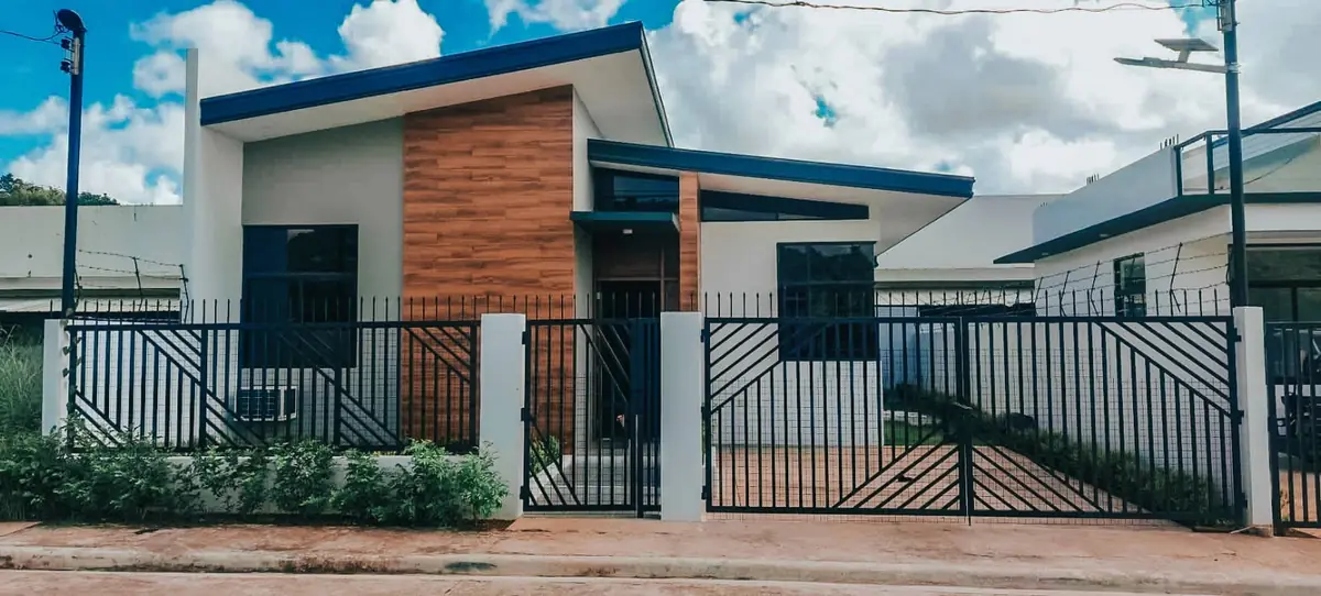 Single storey house with a blue roof and wood grain panel, steel fence and gate on a dirt road