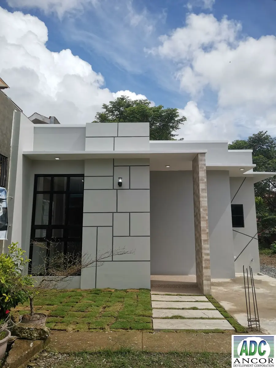 Front exterior of a grey flat roof bungalow with black grid window, covered entry, and grass block paving