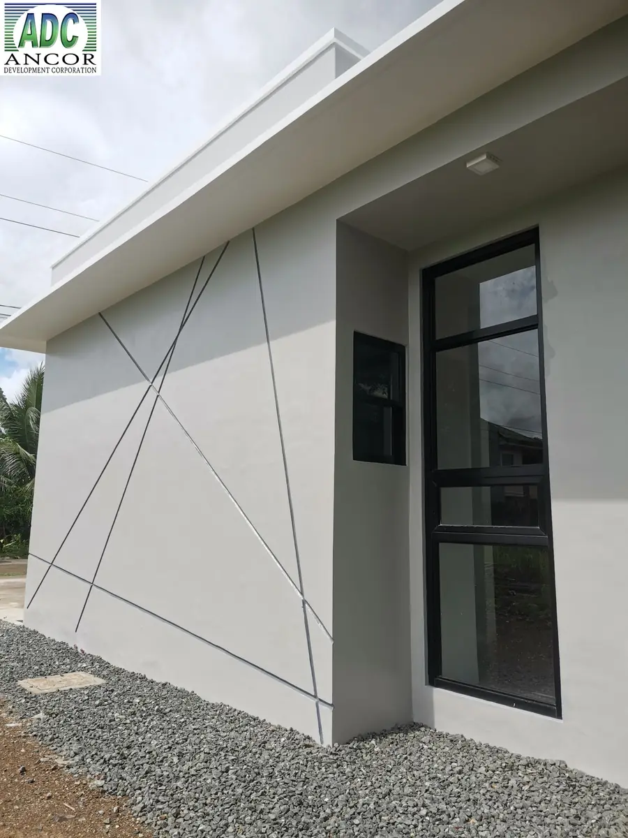 Side exterior wall with diagonal line detail, tall black framed window, and gravel ground