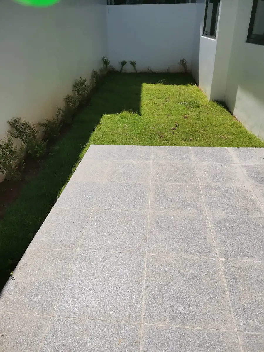 Side yard with lawn, shrub border along white walls, and granite look tiled paving in the foreground