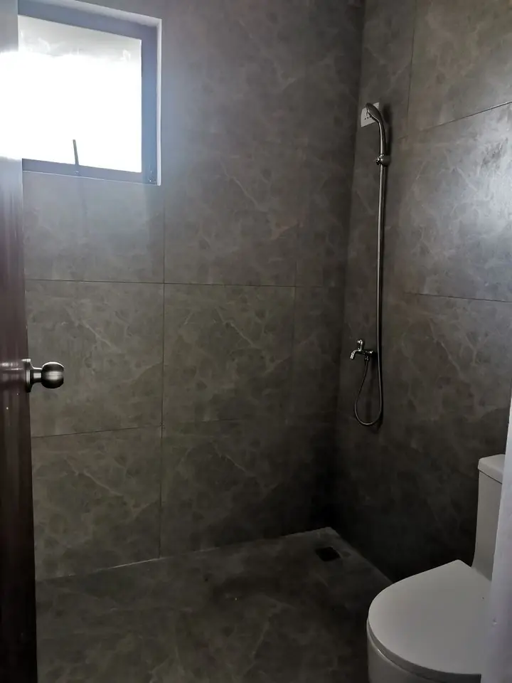 Bathroom with grey tiled walls, wall mounted shower on a rail, floor drain, and a toilet
