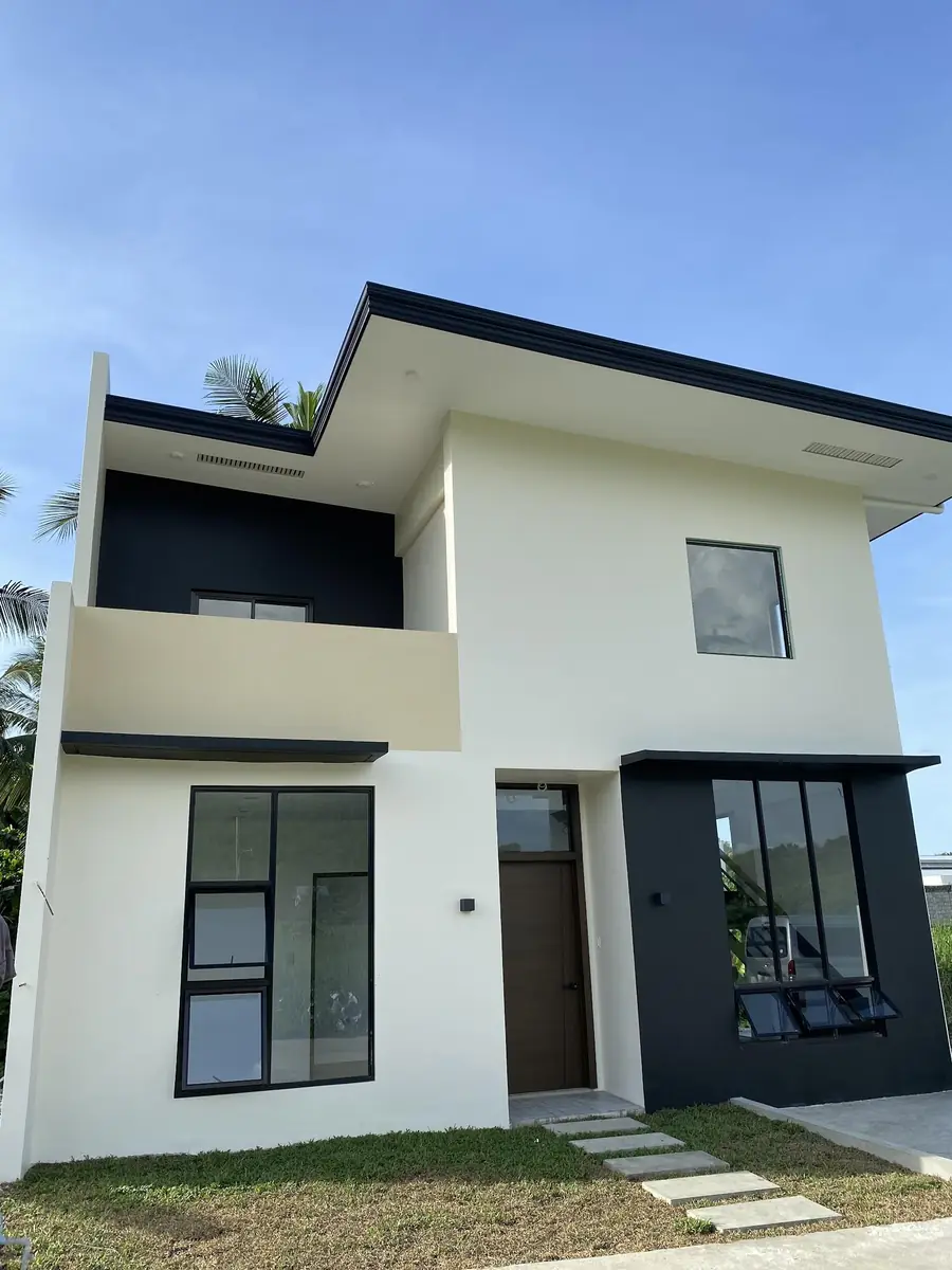 Front of a two storey house with white and dark grey walls, balcony, wood door, and stepping stones