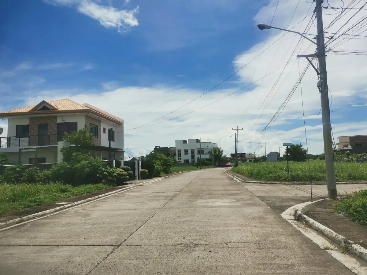 Concrete subdivision road, tan house with orange tiled roof on the left, vacant grass lots on the right