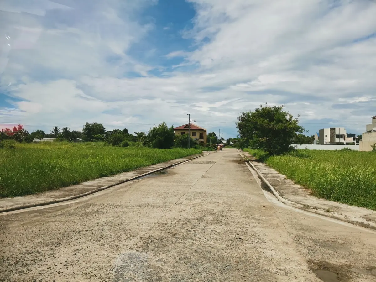 Concrete road between grassy vacant lots, houses and a tricycle in the distance