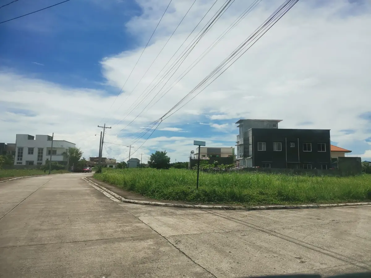 Street corner beside a grassy lot, dark grey three storey building on the right, white houses further off