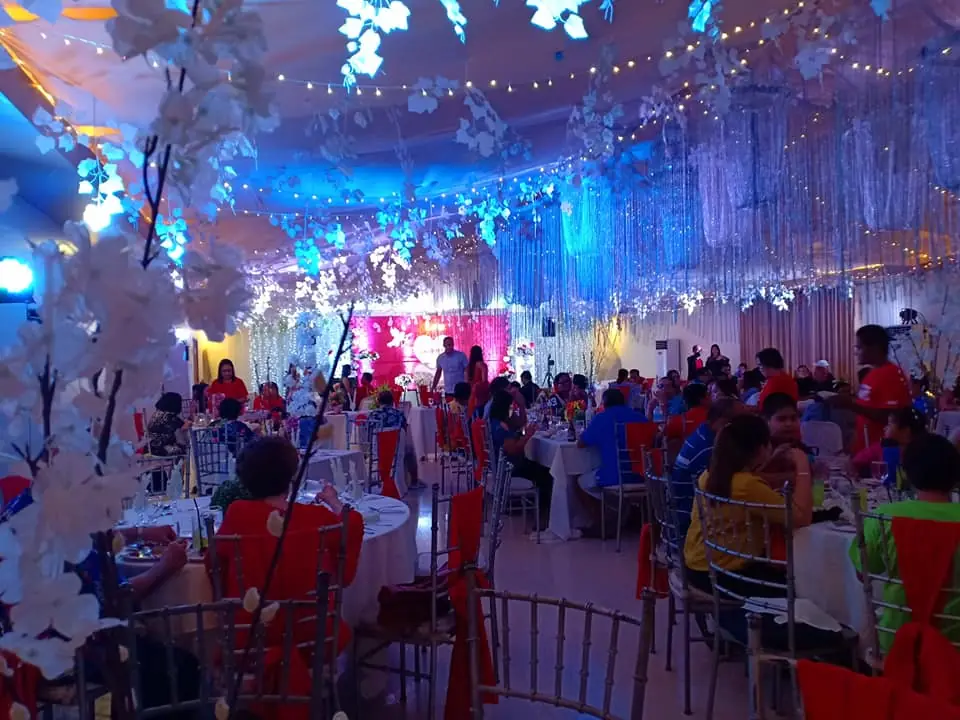 Indoor function hall with round tables, red chair covers, hanging fairy lights and blue uplighting